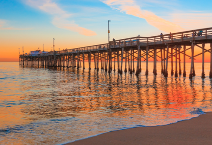 Newport Beach pier in California at sunset over a calm ocean with vibrant orange and pink hues reflecting on the water.